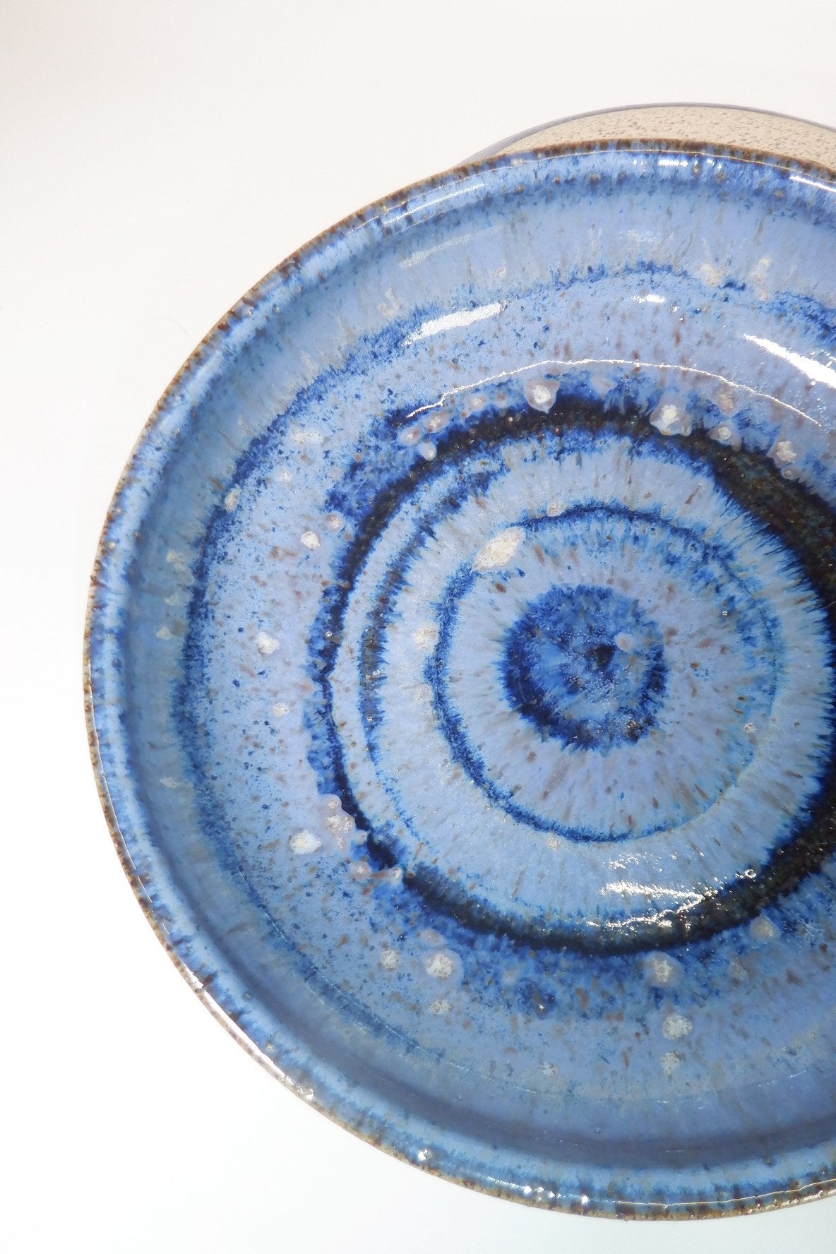 A close-up of a handmade ceramic plate with concentric spiral patterns in shades of blue, from a top-down perspective on a white background.