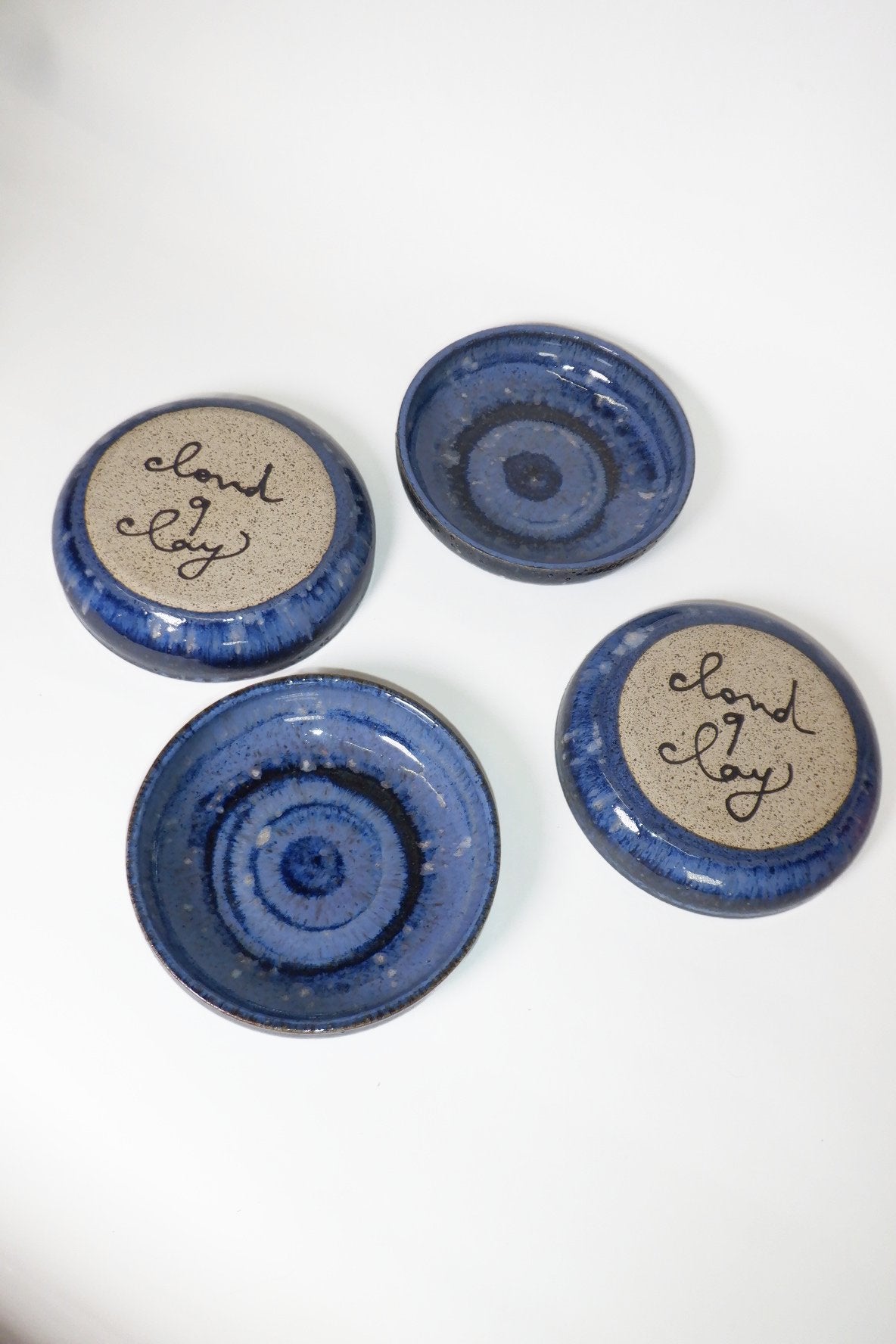 Four small round ceramic dishes in blue and dark blue glaze, two showing the top side with a beige center and the words 'cloud 9 joy' written in black script.