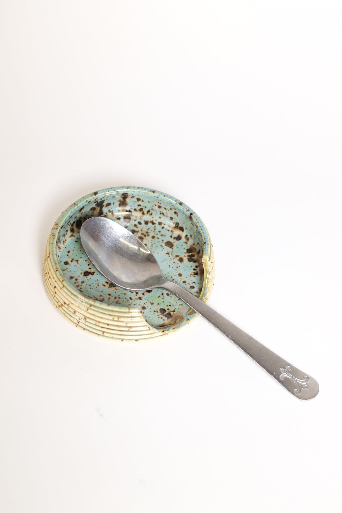A small round ceramic dish with a speckled light blue and white glaze, a gold-toned rim, and a silver metal spoon resting inside it, photographed on a plain white background.