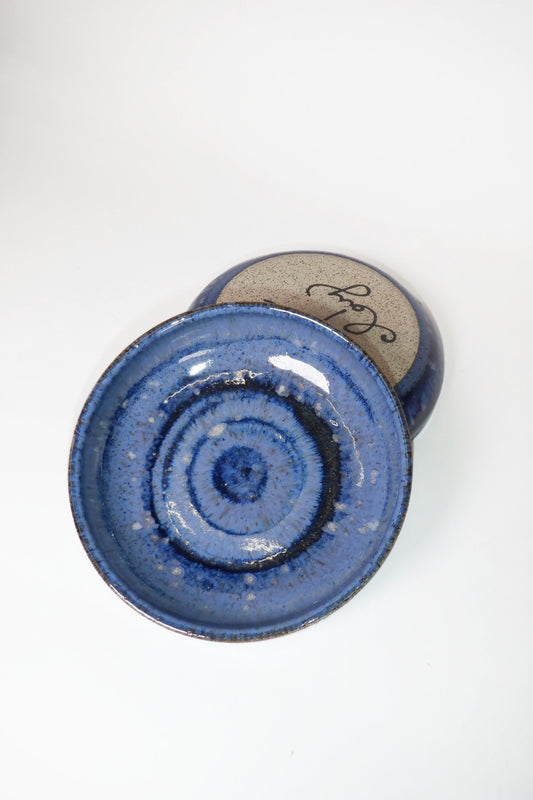 A handmade ceramic bowl with a blue speckled glaze, shown from a top-down angle with its lid resting behind it on a white background.